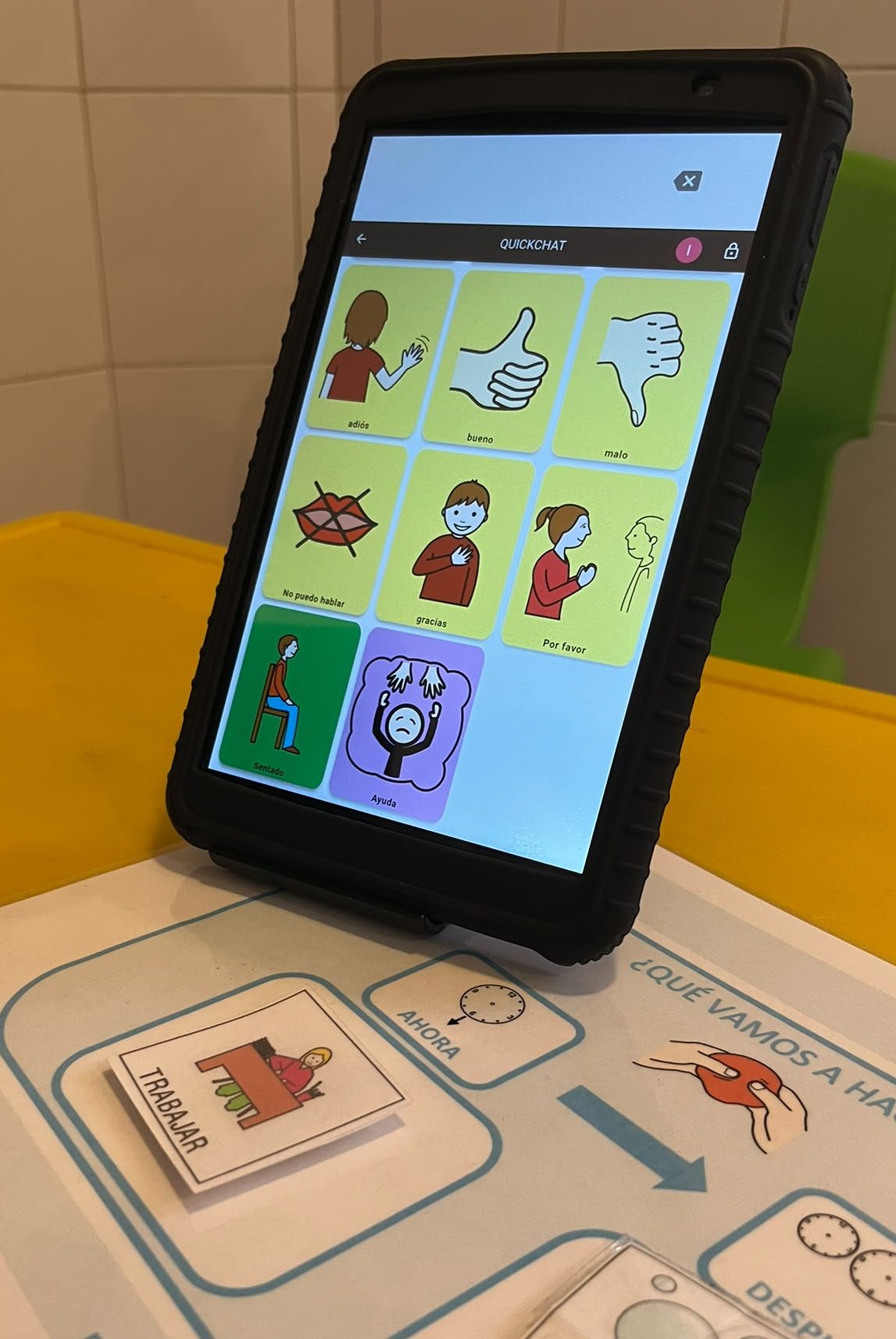Tablet sobre un soporte, mostrando la aplicación de comunicación aumentativa QuickChat con cuadrícula de pictogramas. En la base hay un panel impreso de anticipación visual "¿Qué vamos a hacer?" con las tarjetas de "Ahora: Trabajar" y "Después: Burbujas".